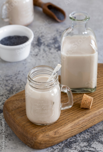 Homemade poppy seed milk in glass jar and milk bottle with scattered seeds on the background. Selective focus, gray concrete background, vertical.