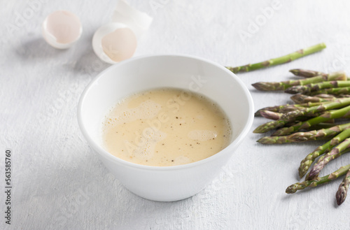 White ceramic bowl of beaten eggs with milk and fresh asparagus shoots on a light gray background. Cooking a delicious homemade omelet