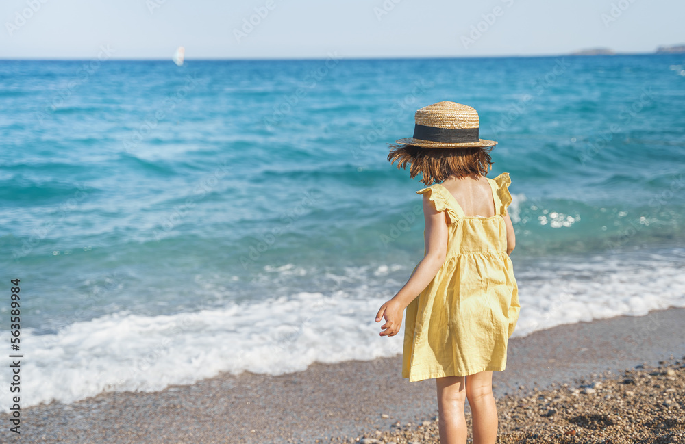 Little girl walking on beach, sea ocean shore in romantic yellow dress, straw hat. Playing in sand, blue waves. Family vacation travel leisure in hot summer coast. Sunny day relax in hotel resort