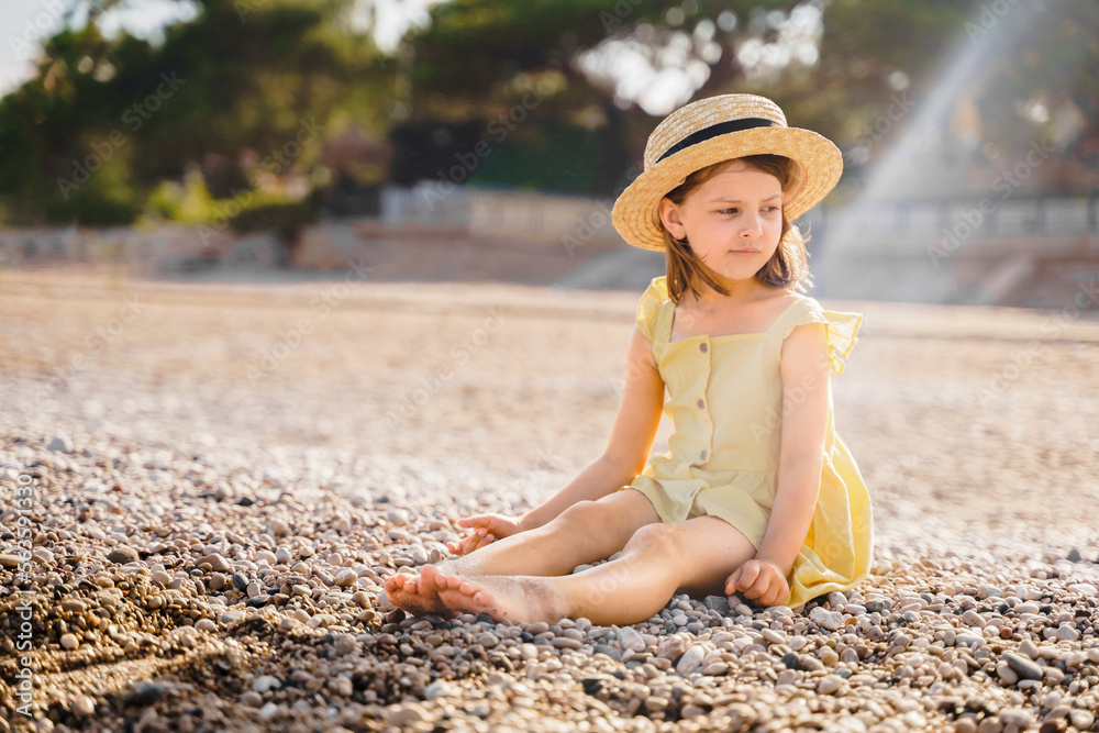 Obraz premium Little girl walking on beach, sea ocean shore in romantic yellow dress, straw hat. Playing in sand, blue waves. Family vacation travel leisure in hot summer coast. Sunny day relax in hotel resort
