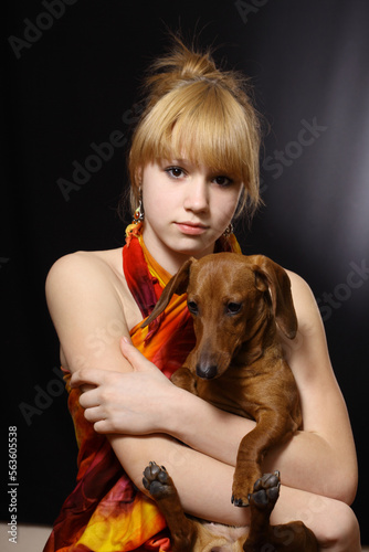 Cute young blonde girl holding a dachshund in her arms isolated on a black background.