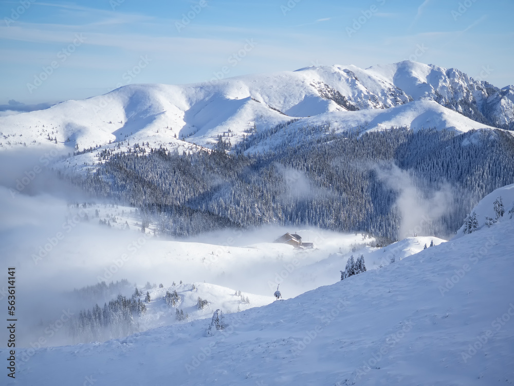 Mountain chalet in winter landscape with low altitude clouds in bright sunny day. Snow covered mountains