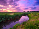 Nature. The river on the field during a bright sunset. Reeds along the river bank. Bright sky with clouds during sunset. Landscape in the summertime. Reflections on the surface of the water.