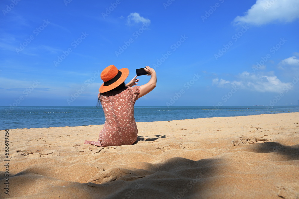 Obraz premium A female tourist wearing a brown skirt and orange hat sits on the sand and uses a mobile phone camera to capture the beauty of the sea and sky on a sunny day. There is a shadow of a tree on the sand. 