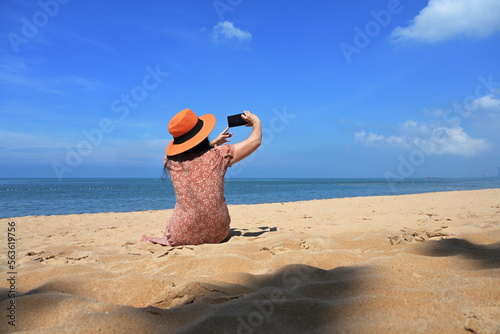 A female tourist wearing a brown skirt and orange hat sits on the sand and uses a mobile phone camera to capture the beauty of the sea and sky on a sunny day. There is a shadow of a tree on the sand. 