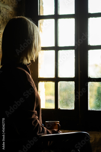 A woman sits near the window with a cup of coffee. Loneliness, sadness concept. Midlife crisis, reflections.