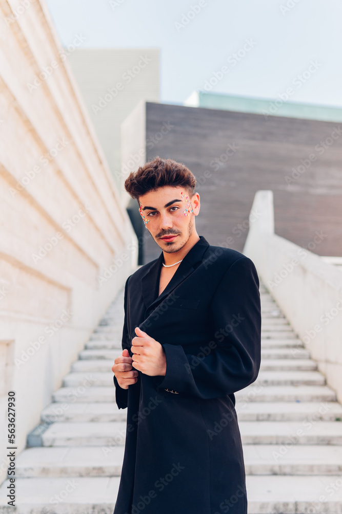Portrait of confident young gay man in long stylish coat standing on stairs looking at camera in city. Confident stylish man in long coat on steps