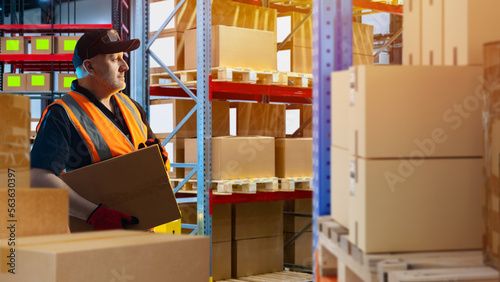 Man with box in warehouse. Worker among stacked racks with boxes. Guy is inside storage building. Worker in reflective vest holds parcel. Sorter in distribution center. Warehouse contractor man photo