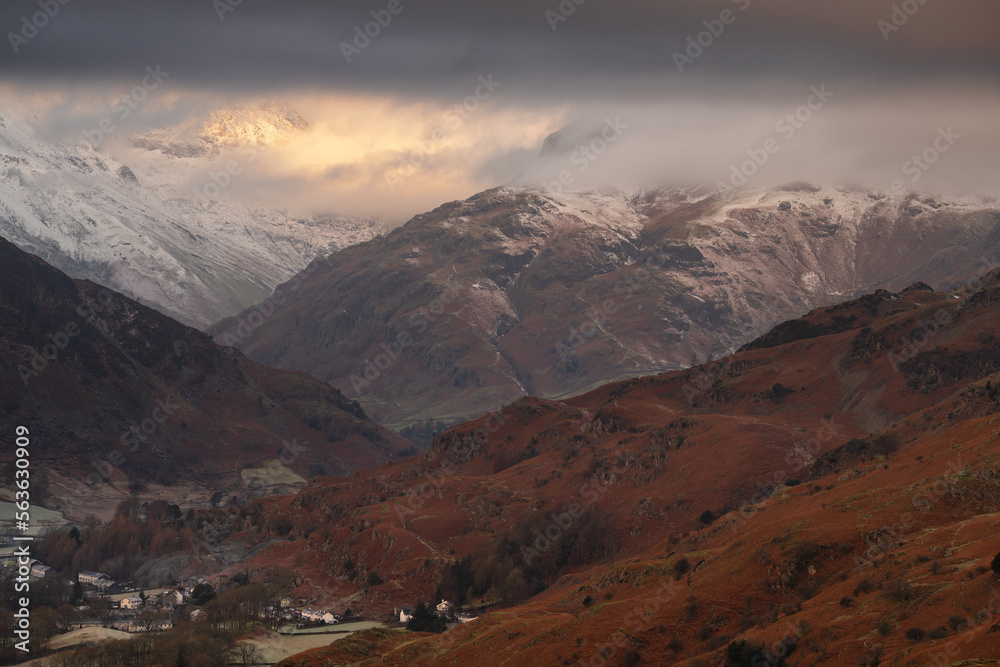 Dramatic view of Lake District snowcapped mountains shrouded in low cloud on a Winter morning with small Cumbrian village; Chapel Stile.