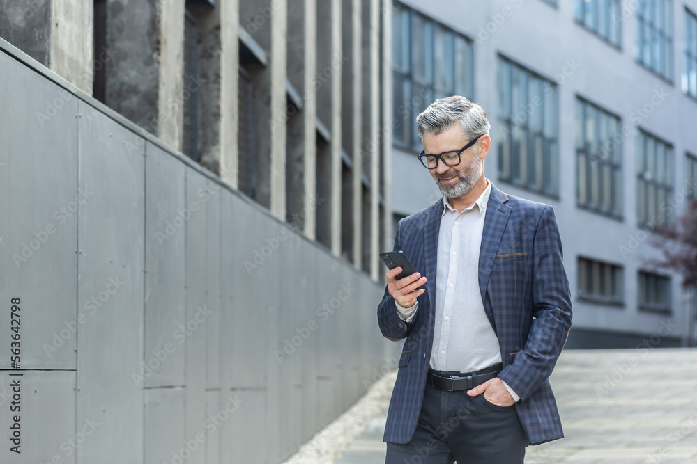 Successful gray haired boss walking outside office building, mature businessman using phone, smiling and reading news typing message, gray haired man in business suit and glasses. Successful gray haired boss walking outside office building, mature businessman using phone, smiling and reading news typing message, gray haired man in business suit and glasses.