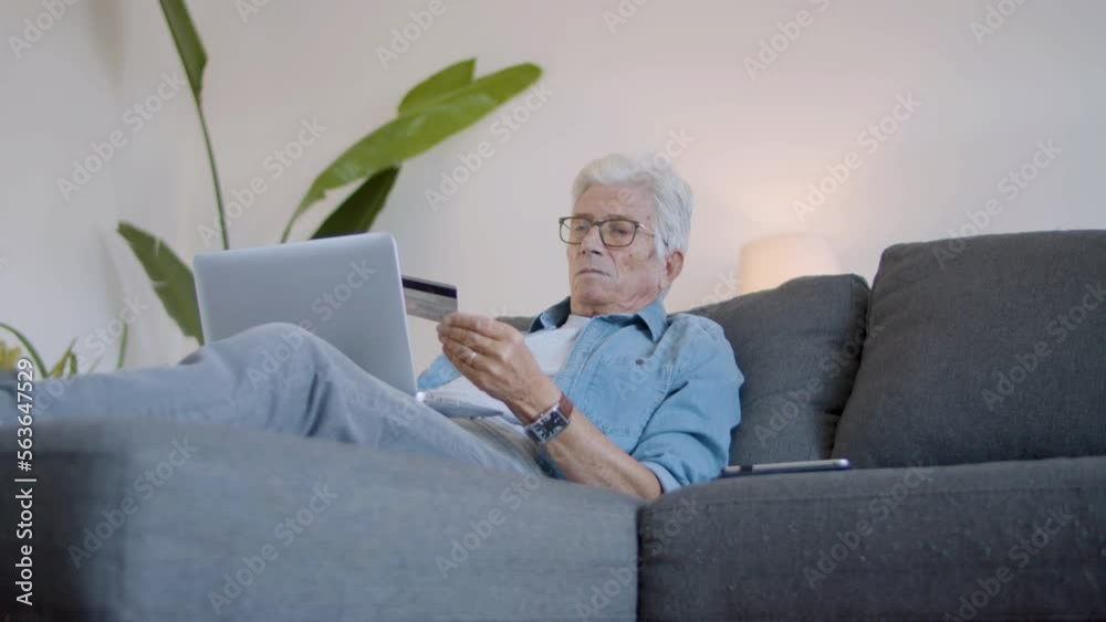 Elderly man doing online shopping on laptop at home. Focused mature man in eyeglasses lying on sofa in living room, entering bank card details to pay for purchase. Digital device, e-commerce concept Elderly man doing online shopping on laptop at home. Focused mature man in eyeglasses lying on sofa in living room, entering bank card details to pay for purchase. Digital device, e-commerce concept