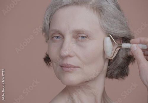 CU Portrait of very beautiful grey-haired 60s mature senior adult female using ceramic face roller. Studio shot, soft lighting. No make up, clean skin