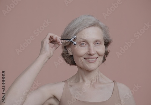 CU Portrait of very beautiful grey-haired 60s mature senior adult female using cosmetics face roller. Studio shot, soft lighting. No make up, clean skin