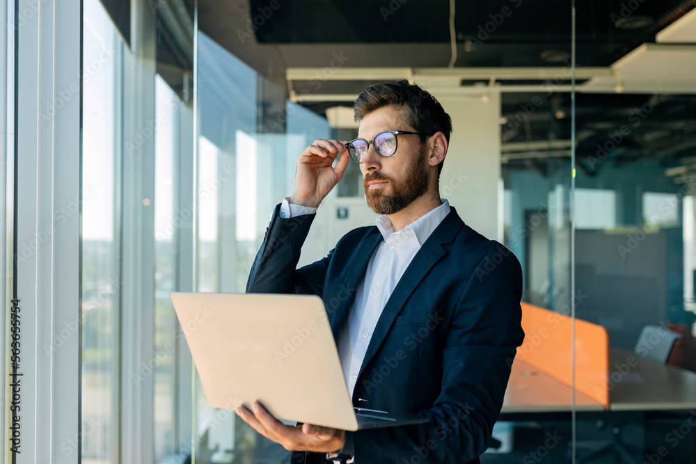 Serious middle aged businessman in glasses holding laptop and looking aside, standing near window in office interior Serious middle aged businessman in glasses holding laptop and looking aside, standing near window in office interior