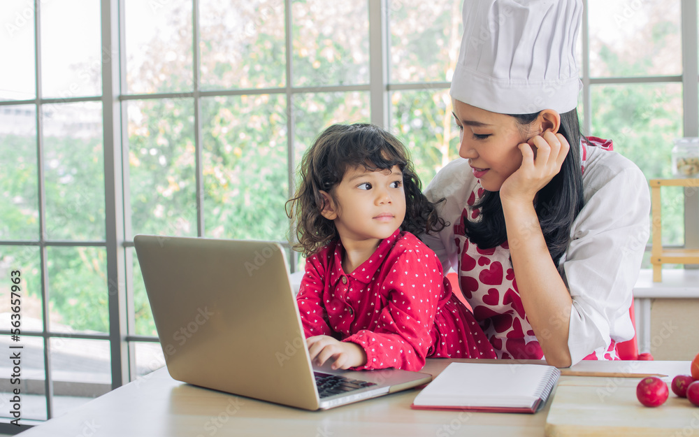 Beautiful Asian mother cooking for breakfast while taking care her mixed race adorable little daughter to do homework, online study or learning, sitting in kitchen at home, smiling with happiness. Beautiful Asian mother cooking for breakfast while taking care her mixed race adorable little daughter to do homework, online study or learning, sitting in kitchen at home, smiling with happiness.