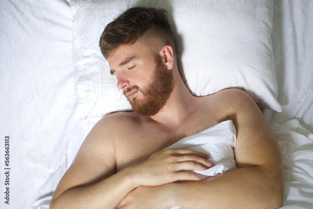 Handsome young calm sleepy bearded man with beard is sleeping well on side in bed in bedroom on pillow with his eyes closed in the morning, resting. Good healthy dream, rest. White linens. Top view. Handsome young calm sleepy bearded man with beard is sleeping well on side in bed in bedroom on pillow with his eyes closed in the morning, resting. Good healthy dream, rest. White linens. Top view.