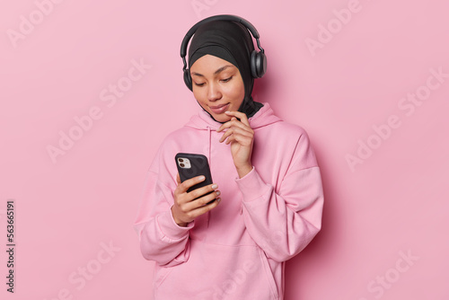 Horizontal shot of Muslim woman focused attentively at screen of smartphone chooses song to listen from playlist uses stereo headphones wears hijab and sweatshirt isolated over pink background
