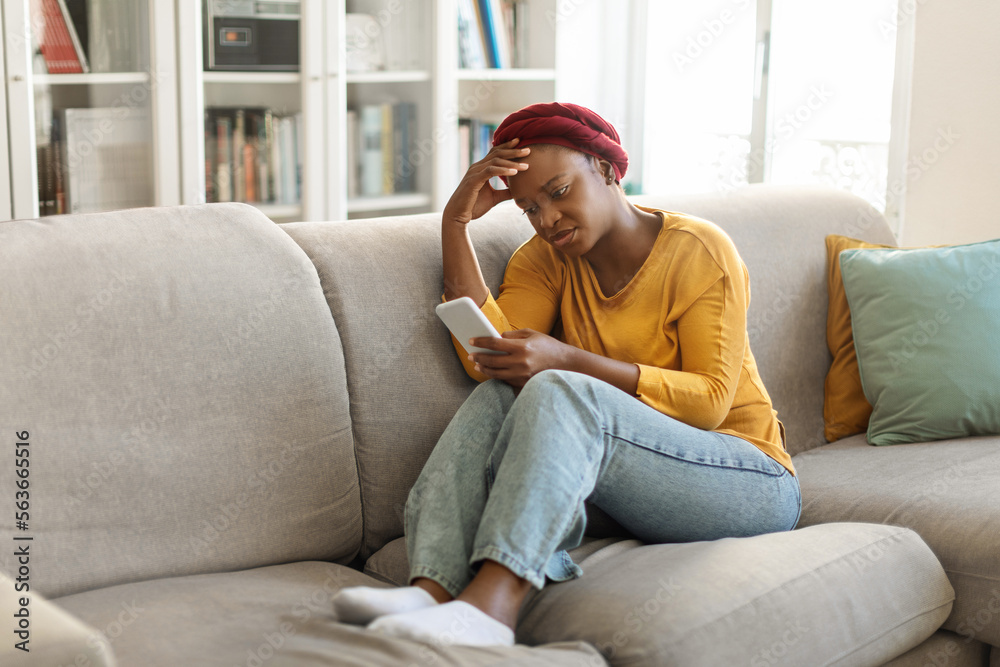 Upset black woman sitting on sofa with cellphone, home interior Upset black woman sitting on sofa with cellphone, home interior