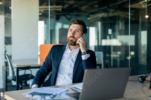Confident middle aged businessman calling by cellphone, sitting at workplace with laptop and communicating in office