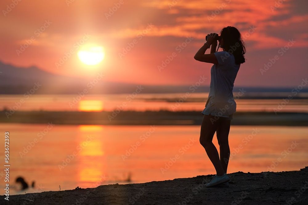 On safari in Zimbabwe: Silhouette of a Fit Woman standing on the Banks of the Zambezi River, observing African nature through binoculars, against red setting sun reflecting on water surface.