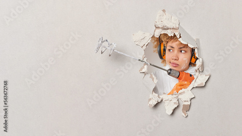 Serious female builder does renovation in her house holds paint mixer in hands breaks through white wall uses headphones for noise cancellation focused aside. Redecoration and repair concept.