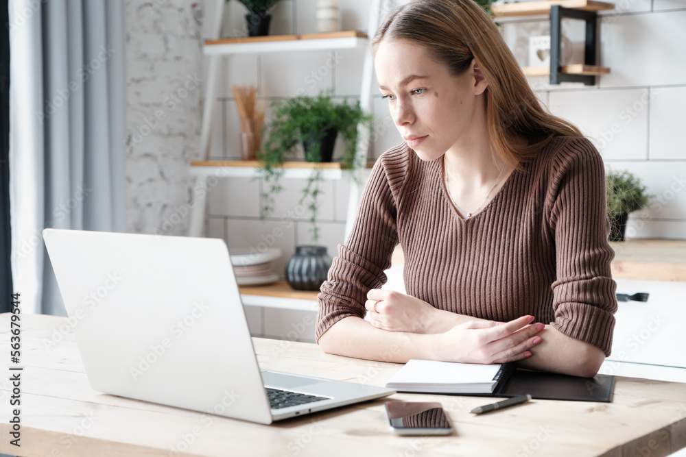 Thoughtful caucasian young female remote worker in casual outfit sitting at table at home with laptop, smartphone and planner doing work task Thoughtful caucasian young female remote worker in casual outfit sitting at table at home with laptop, smartphone and planner doing work task