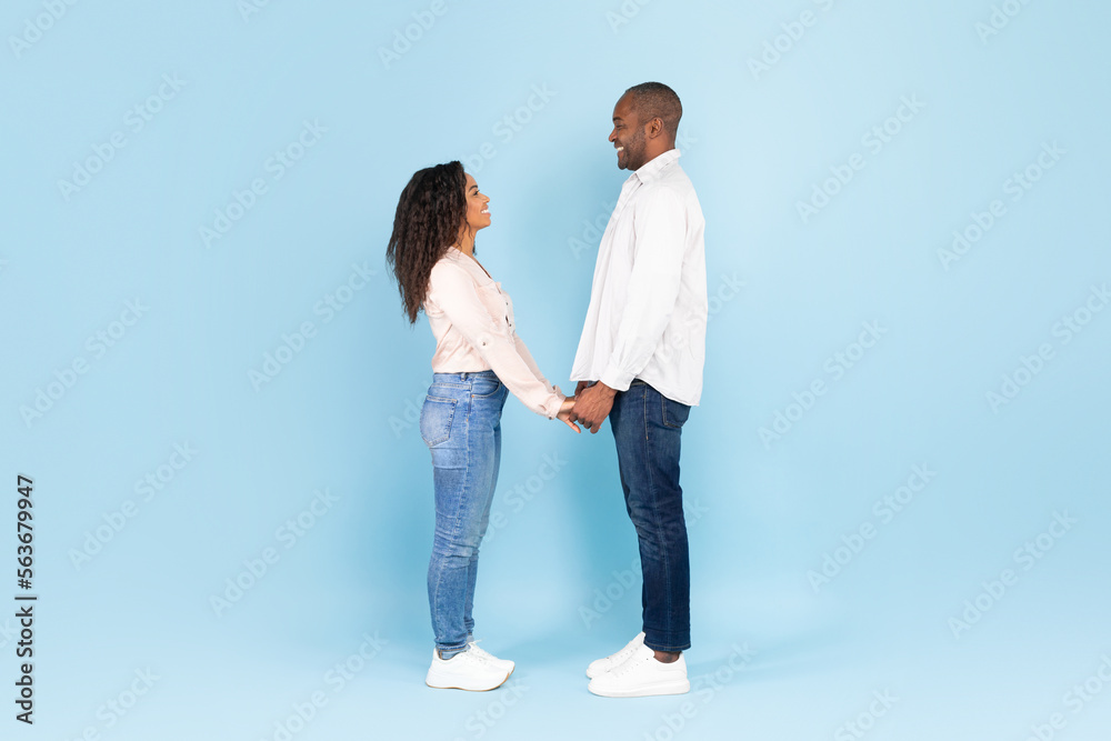 Full length of black couple holding hands and looking at each other against blue background, side view, free space Full length of black couple holding hands and looking at each other against blue background, side view, free space