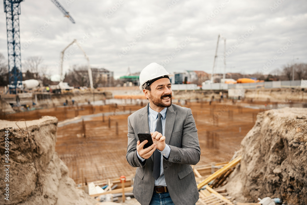 A happy engineer is standing on construction site with building foundation and calling client on the phone.