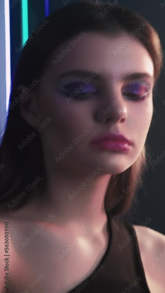 Vertical video. 90s model. Retro fashion. Old 2000s look. Portrait of confident teen girl face with artistic makeup posing in purple blue color neon light on dark black.