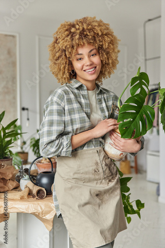 Happy female florist gives tips about easy houseplants care holds potted flower going to transplant into larger container fertilizes or propagates it wears checkered shirt and apron uses fresh soil