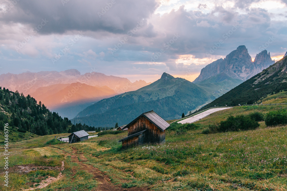 Traditional wooden houses in the Italian Alps in a cinematic landscape with golden light, a mountain road and impressive mountains. Dolomites, Italy. Popular travel destination. Summer tourism.