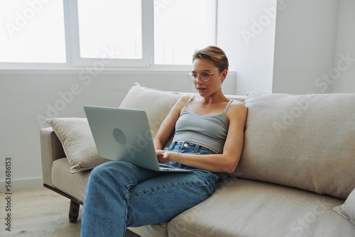 Teenage girl freelancer with laptop sitting on couch at home smiling in home clothes and glasses with short haircut, lifestyle with no filters, free copy space