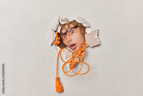 Stunned speechless woman stares throguh transparent glasses keeps mouth opened from wonder busy doing reconstruction and repair uses electric extension cord breaks plaster wall. Renovation concept photo