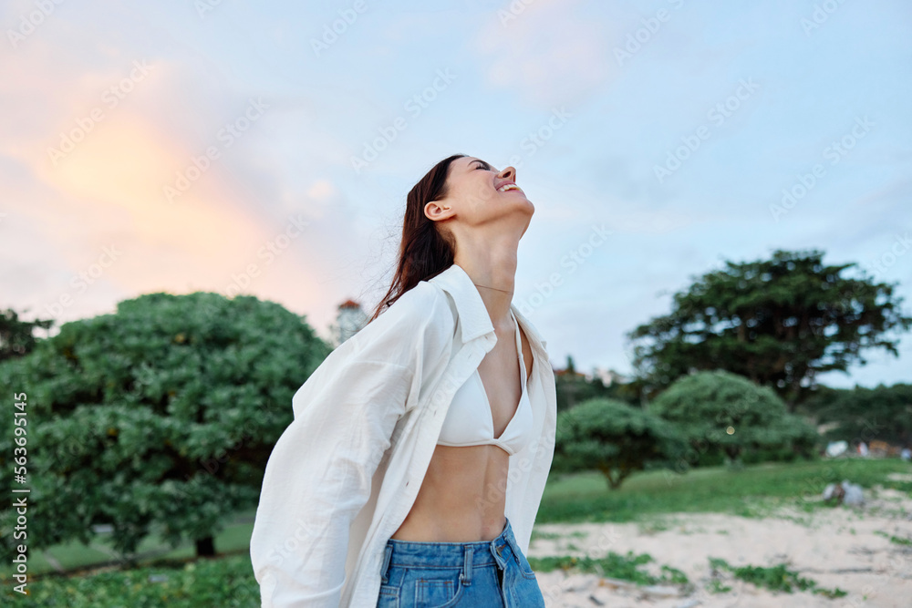 Brunette woman with long hair in a white shirt and jean shorts tan and happy fun smile with teeth in the background of the beach and palm trees, vacation summer trip sunset sky