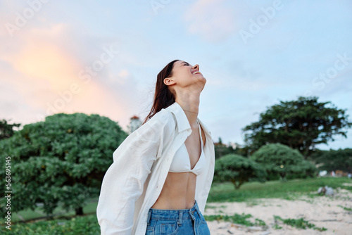 Brunette woman with long hair in a white shirt and jean shorts tan and happy fun smile with teeth in the background of the beach and palm trees, vacation summer trip sunset sky © SHOTPRIME STUDIO