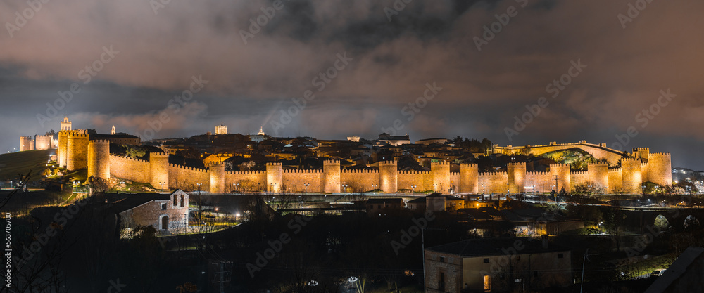 Panoramic view of the old town of Ávila, Spain, surrounded by a medieval wall, lit up during a cold cloudy winter night.