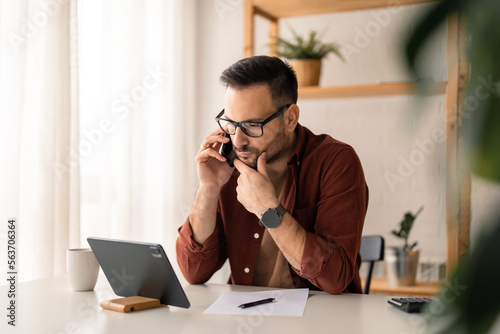 Busy handsome young businessman, broker or advisor having conversation on smart phone from home office, consulting client, looking at digital tablet thinking on financial data investment.