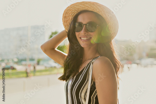 Close up outdoor photo of charming beautiful woman with happy smile wearing straw hat and sunglasses posing at camera in sunlight on background of sunny city