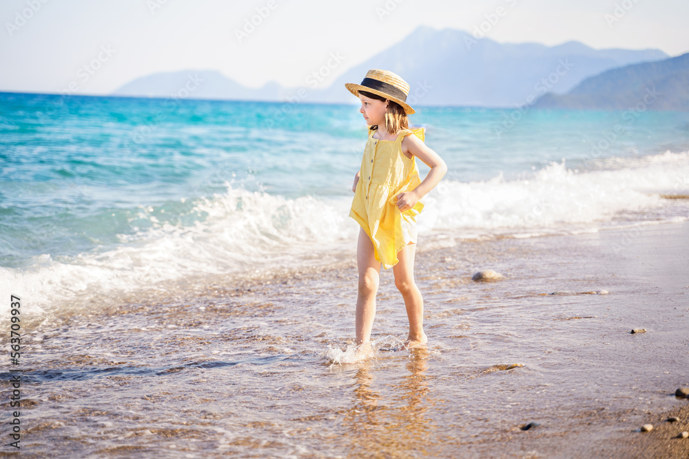 Little girl walking on beach, sea ocean shore in romantic yellow dress, straw hat. Playing in sand, blue waves. Family vacation travel leisure in hot summer coast. Sunny day relax in hotel resort Little girl walking on beach, sea ocean shore in romantic yellow dress, straw hat. Playing in sand, blue waves. Family vacation travel leisure in hot summer coast. Sunny day relax in hotel resort
