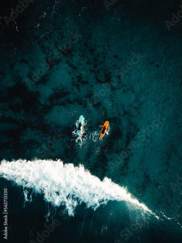 Two surfers floating on surfboard in tropical ocean with big and powerful wave behind them. Aerial top view of surfers in crystal blue ocean - Vertical photo.
