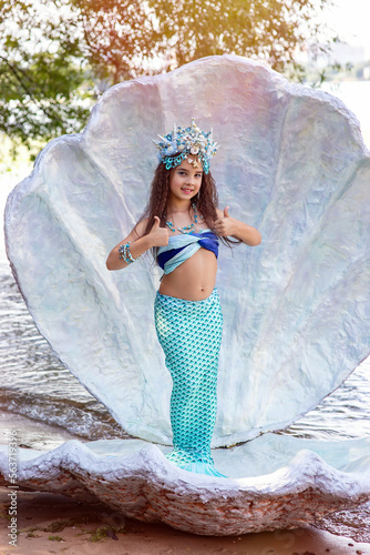 A beautiful girl in a mermaid costume stands outdoor in a large sea shell, holds her thumbs up