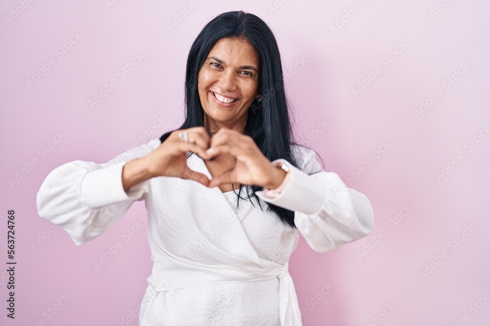 Mature hispanic woman standing over pink background smiling in love doing heart symbol shape with hands. romantic concept.