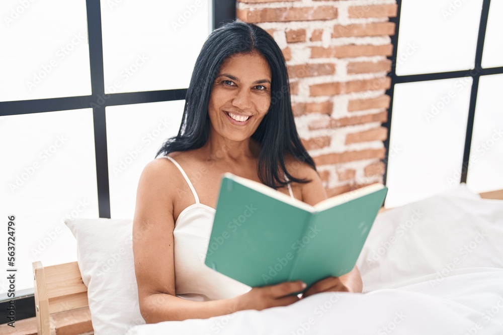 Middle age hispanic woman reading book sitting on bed at bedroom Middle age hispanic woman reading book sitting on bed at bedroom