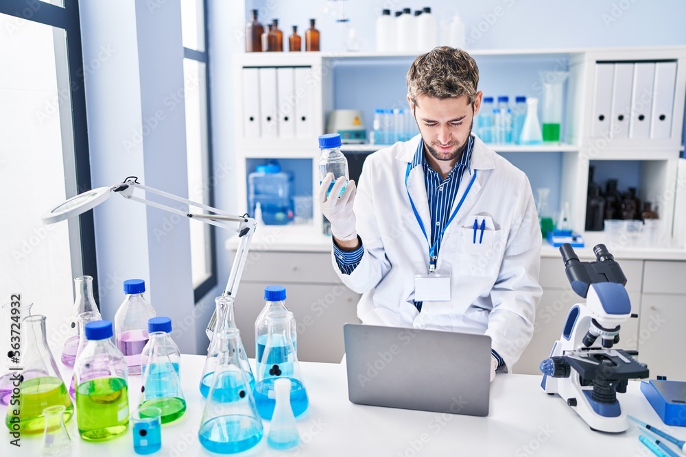 Young man scientist using laptop holding bottle at laboratory