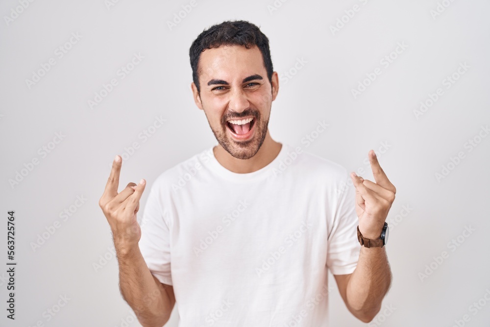Handsome hispanic man standing over white background shouting with crazy expression doing rock symbol with hands up. music star. heavy music concept. Handsome hispanic man standing over white background shouting with crazy expression doing rock symbol with hands up. music star. heavy music concept.