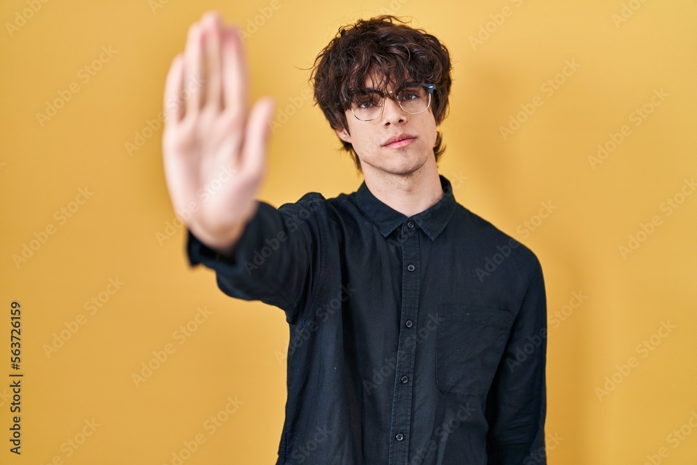 Young man wearing glasses over yellow background doing stop sing with palm of the hand. warning expression with negative and serious gesture on the face.