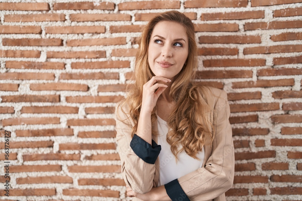 Beautiful blonde woman standing over bricks wall with hand on chin thinking about question, pensive expression. smiling with thoughtful face. doubt concept.