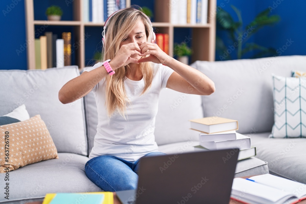 Young blonde woman studying using computer laptop at home doing heart shape with hand and fingers smiling looking through sign Young blonde woman studying using computer laptop at home doing heart shape with hand and fingers smiling looking through sign