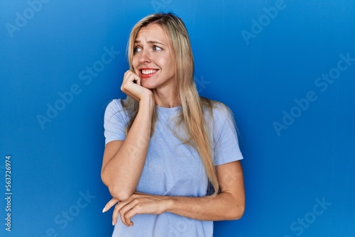 Beautiful blonde woman wearing casual t shirt over blue background looking stressed and nervous with hands on mouth biting nails. anxiety problem.