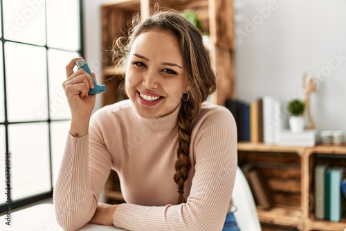 Young beautiful hispanic woman using inhaler sitting on table at home